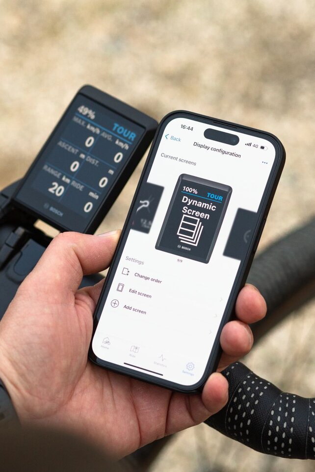 Une personne tient un smartphone affichant l’application Bosch eBike FlowApp avec la page « Configuration de l'écran » et l'« écran/page dynamique ». Le smartphone est placé à côté d’un guidon de vélo équipé d’un écran numérique qui affiche des informations telles que le niveau de la batterie, le mode (TOUR), la vitesse et l'autonomie. L’arrière-plan est flou et montre un environnement extérieur.