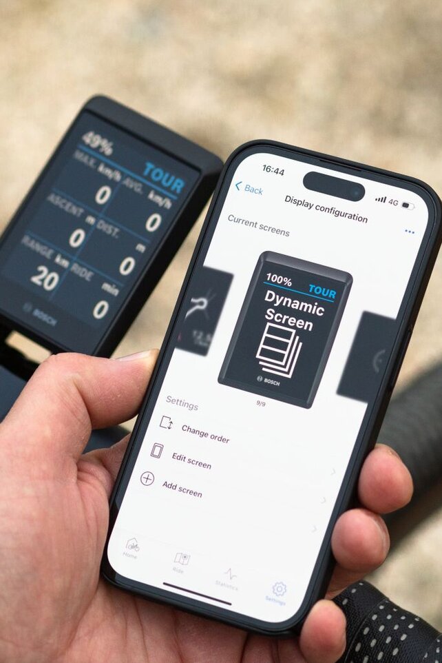 Una persona sostiene un teléfono inteligente que muestra la Bosch eBike FlowApp con la indicación "Configuración de display" y "pantalla dinámica". El teléfono se sostiene junto a un manillar de bicicleta equipado con un display digital que muestra información como el nivel de batería, el modo (TOUR), velocidad y autonomía. El fondo está desenfocado y muestra un entorno al aire libre.