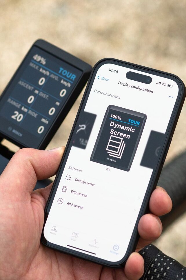 A person is holding a smartphone displaying the Bosch eBike FlowApp with the screen layout "display configuration" and "Dynamic screen". The smartphone is held next to a bicycle handlebar equipped with a digital display that shows information such as battery level, mode (TOUR), speed, and range. The background is blurred and reveals an outdoor environment.