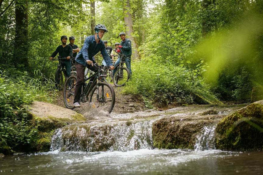 eBike-ryttere på en sti i den grønne skogen, rett før de skal krysse en bekk.