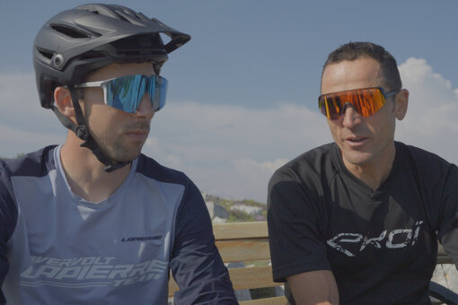 Deux hommes en tenue de VTT sont assis côte à côte sur un banc. À l’arrière-plan, on aperçoit des pneus de vélo et un ciel bleu légèrement nuageux.
