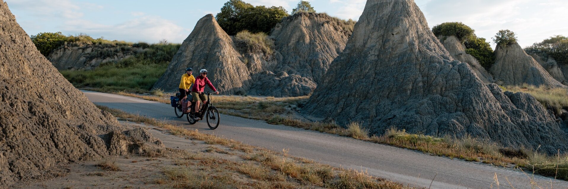 Two people are riding e-bikes along a road through a rocky landscape with striking rock formations. One person is wearing a yellow jacket, the other a red jacket. Both bikes have panniers attached. The sky is partly cloudy.