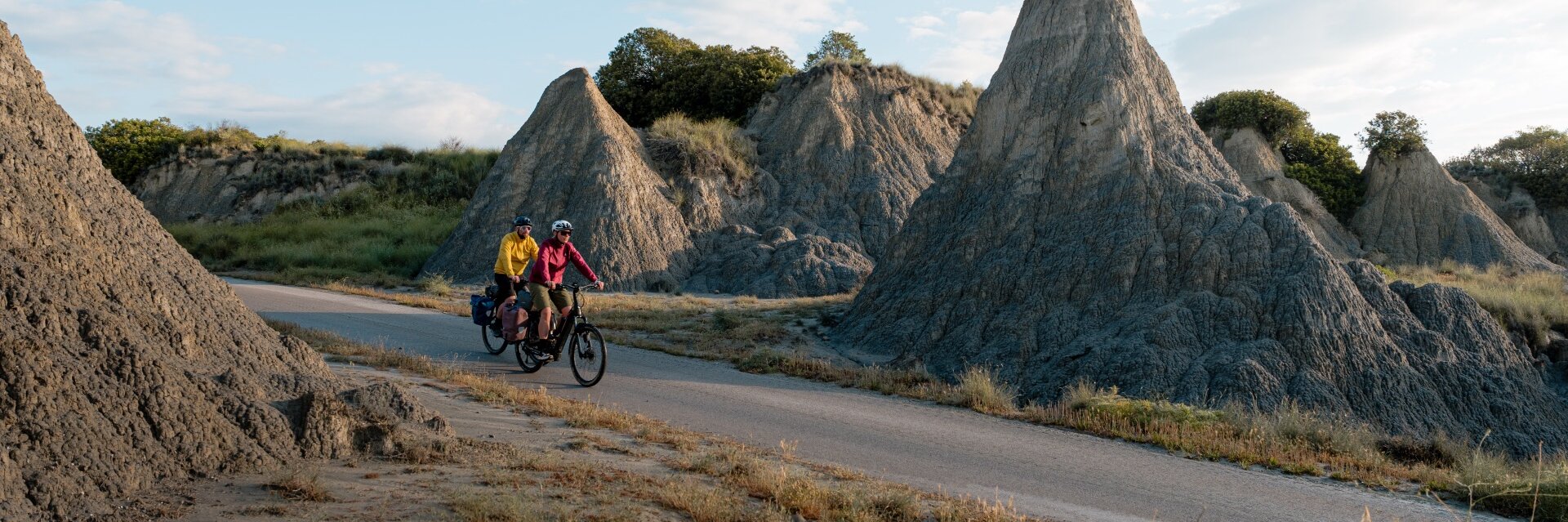  Zwei Personen fahren mit E-Bikes auf einer Straße durch eine felsige Landschaft mit ungewöhnlichen Felsformationen. Eine Person trägt eine gelbe Jacke, die andere eine rote Jacke. Beide Fahrräder sind mit Gepäcktaschen ausgestattet. Der Himmel ist leicht bewölkt.