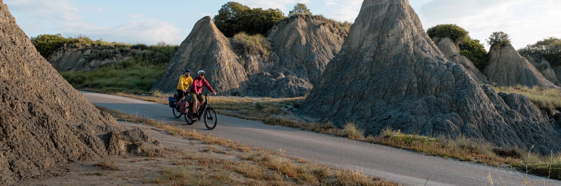 Two people are riding e-bikes along a road through a rocky landscape with striking rock formations. One person is wearing a yellow jacket, the other a red jacket. Both bikes have panniers attached. The sky is partly cloudy.