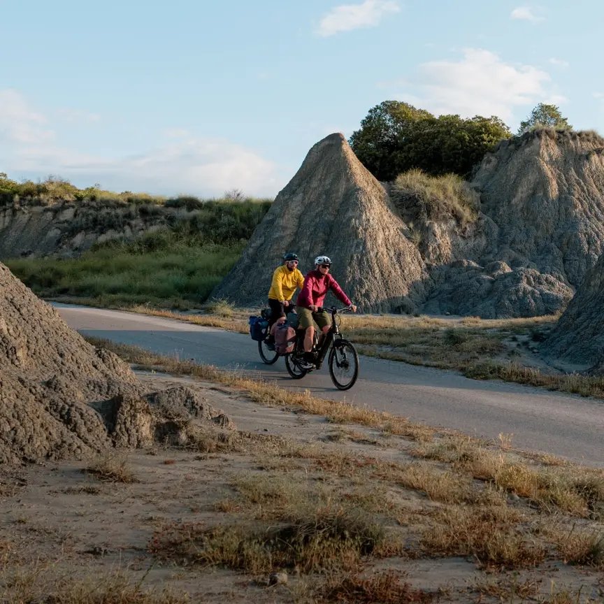 Two people are riding e-bikes along a road through a rocky landscape with striking rock formations. One person is wearing a yellow jacket, the other a red jacket. Both bikes have panniers attached. The sky is partly cloudy.