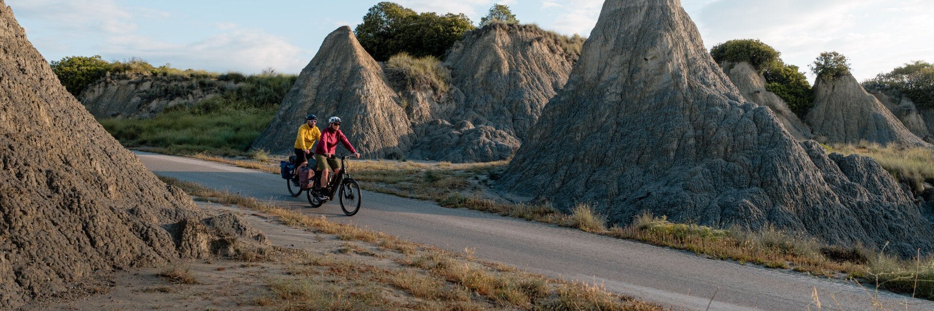 Due persone viaggiano in eBike lungo una strada che attraversa un paesaggio roccioso con formazioni rocciose particolari. Una persona indossa una giacca gialla, l'altra una giacca rossa. Entrambe le eBike sono equipaggiate con borse laterali. Il cielo è leggermente nuvoloso.