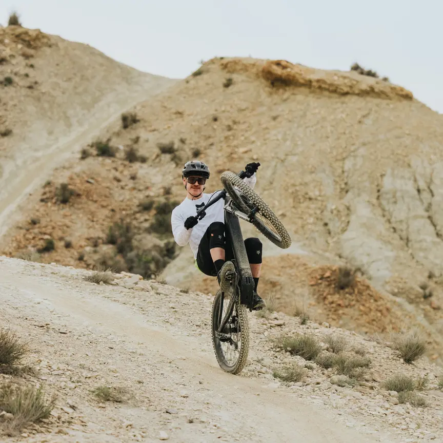 En eMountainbiker i fuldt udstyr smiler, mens han laver en wheelie på baghjulet og kører ad en grussti gennem et tørt, kuperet landskab.