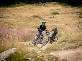 Two mountain bikers are riding up a narrow trail and leaning into the curve.