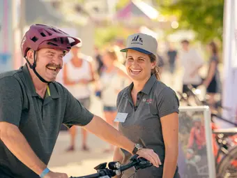 A man is sitting on an eBike, laughing, with a Bosch specialist standing next to him, also laughing. In the background, more people and bicycles can be seen.