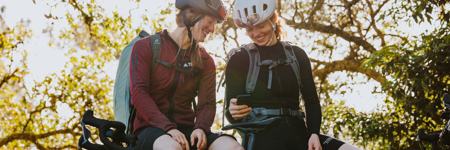 Two women sit on a wall, looking at a smartphone and wearing bicycle helmets. The sun is shining on trees behind them, and their eBikes are in front of them.