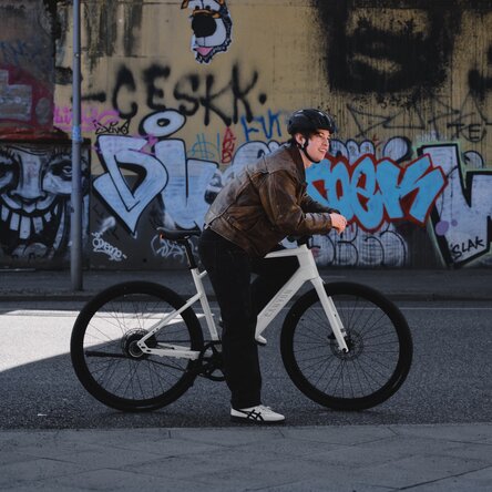 Un joven ciclista con casco y chaqueta de cuero se ha detenido y se apoya tranquilamente en su eBike blanca. Está de pie, sonriente, frente a un muro totalmente cubierto de pintadas coloridas, lo que aporta a la escena un marcado carácter urbano.