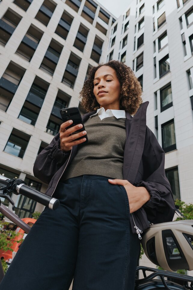 Shot of a woman from a low angle. She holds her smartphone and looks at it; her eBike stands in the background, and her helmet hangs casually over her arm.