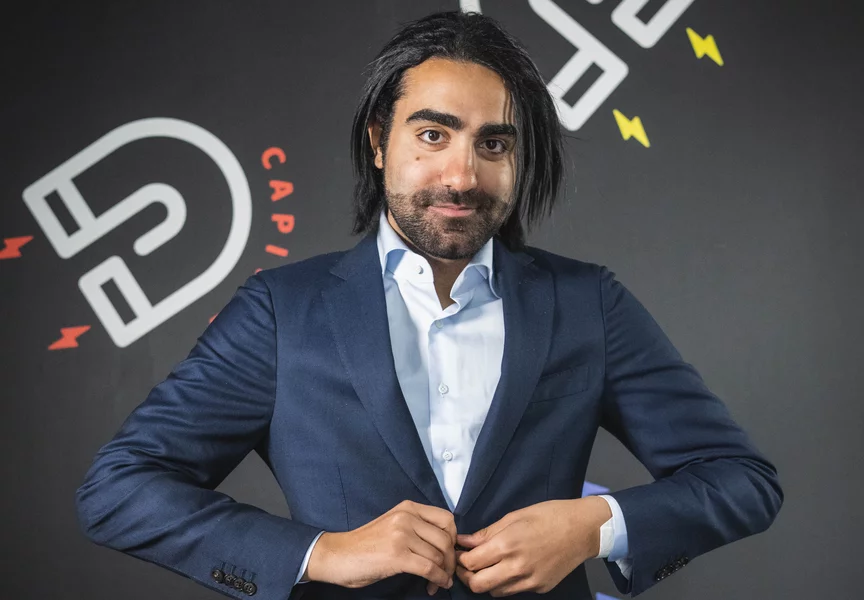 Portret van een man voor een muur, die zelfverzekerd in de camera kijkt terwijl hij de knoop van zijn colbert dichtdoet.
