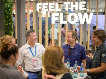 Claus Fleischer addresses a small group at a standing table in front of a Bosch exhibition stand in high spirits. In the background, the illuminated sign "Feel the Flow" is visible.
