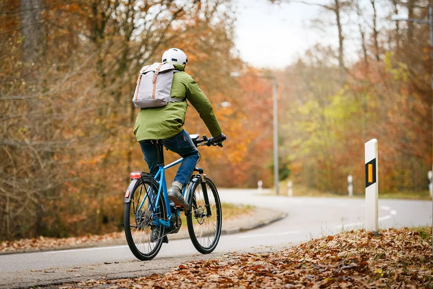 Een eBike-rijder in een groene jas rijdt op een blauwe eBike langs een weg. De weg is omzoomd door bomen met kleurrijke bladeren, waar al bladeren op de grond liggen.