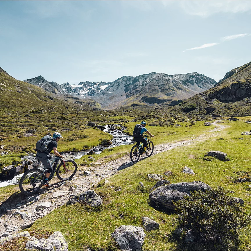 Dos ciclistas en eBike recorren un camino a lo largo de un arroyo en las montañas en terreno llano.