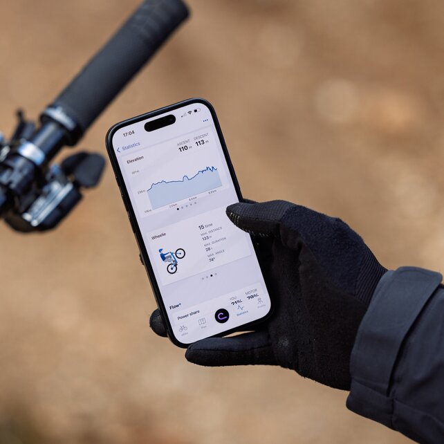 Nahaufnahme einer behandschuhten Hand, die ein Smartphone über einem eBike-Lenker hält. Auf dem Display wird in der eBike Flow App die Statistik einer eBike-Tour angezeigt, inklusive Höhenprofil und Trick-Statistik der absolvierten Wheelies.