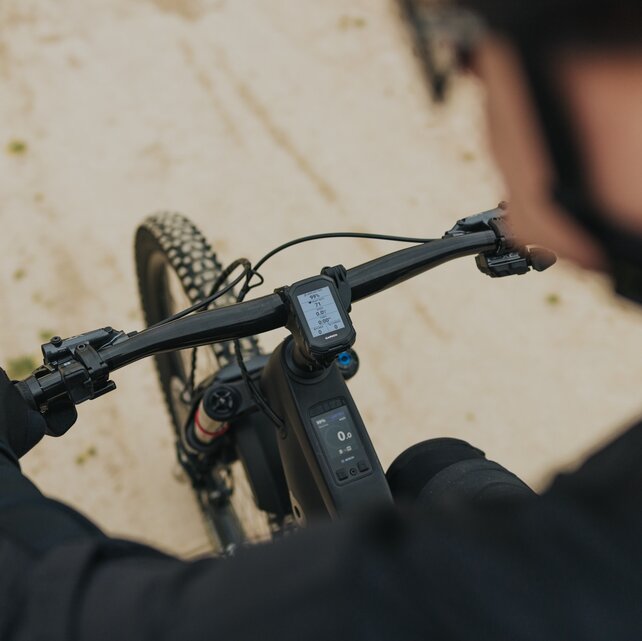 Aufnahme aus der Fahrerperspektive, die über die Schulter auf das Cockpit eines eMountainbikes blickt. Die behandschuhten Hände liegen am Lenker, während der Blick auf die beiden Displays – einen Garmin-Fahrradcomputer und das integrierte Kiox 400C von Bosch im Oberrohr – sowie den sandigen Weg vor dem Rad fällt.