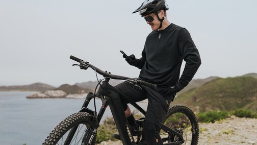 Un Vététiste est assis sur son VAE et fait une pause pour consulter son smartphone. Il se trouve sur un belvédère offrant une vue sur une baie pittoresque et un village côtier en arrière-plan.