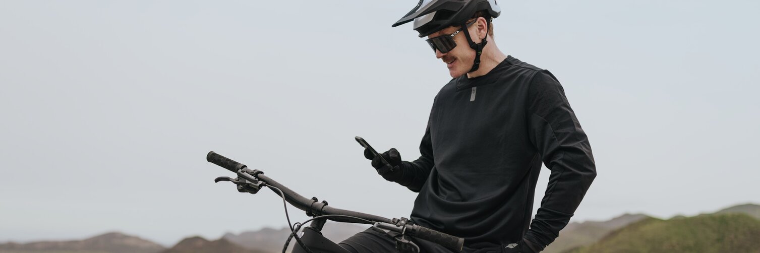 An eMountain biker sits on his eBike and takes a break to check his smartphone. He’s at a viewpoint overlooking a picturesque bay with a coastal village in the background.
