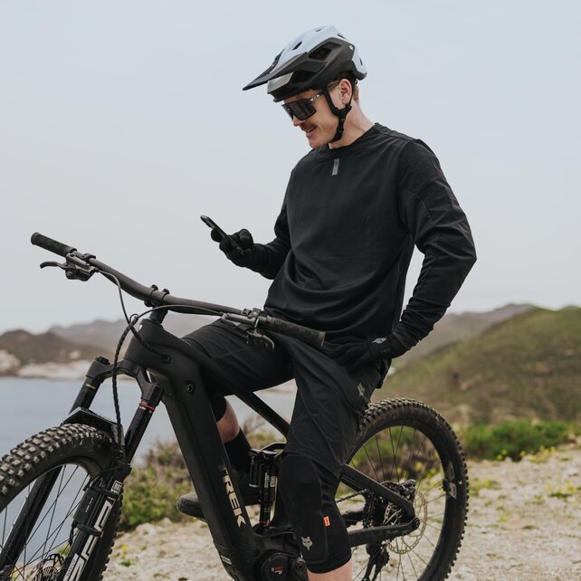 Ein eMountainbiker sitzt auf seinem eBike und macht eine Pause, um auf sein Smartphone zu schauen. Er befindet sich auf einem Aussichtspunkt mit Blick auf eine malerische Bucht und ein Küstendorf im Hintergrund.