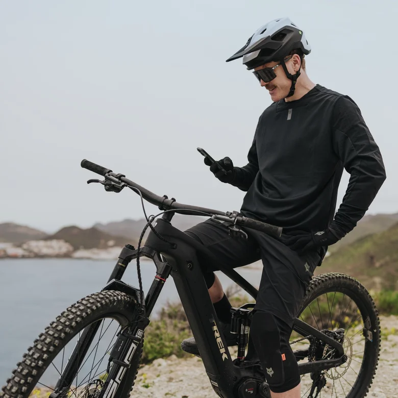 An eMountain biker sits on his eBike and takes a break to check his smartphone. He’s at a viewpoint overlooking a picturesque bay with a coastal village in the background.