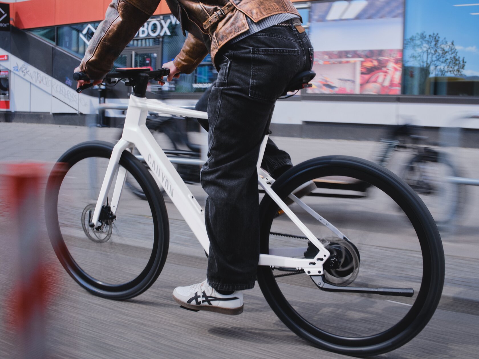 Primer plano de una persona conduciendo un moderno eBike blanco con un motor de cubo en un entorno urbano. La cámara sigue el movimiento, difuminando el fondo con las fachadas de las tiendas y creando una fuerte sensación de velocidad.