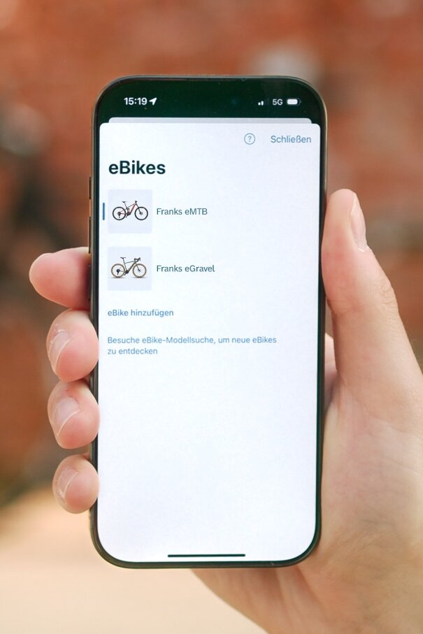 Nahaufnahme eines Smartphones, das die digitale Garage der eBike Flow App anzeigt. Auf dem Bildschirm ist eine Liste mit den persönlichen eBikes des Nutzers zu sehen, darunter ein eMTB und ein eGravel Bike, sowie die Option, weitere eBikes hinzuzufügen.