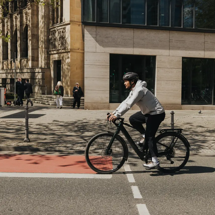 A young man in casual clothing and wearing a bicycle helmet rides an eBike on a city street, crossing a red bike path. Modern buildings and passersby are visible in the background.