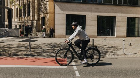 A young man in casual clothing and wearing a bicycle helmet rides an eBike on a city street, crossing a red bike path. Modern buildings and passersby are visible in the background.