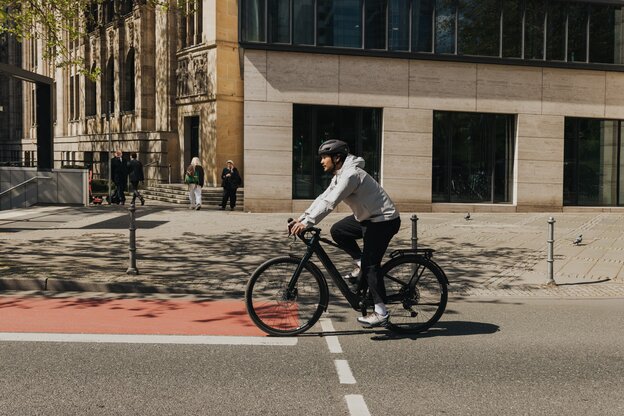 A young man in casual clothing and wearing a bicycle helmet rides an eBike on a city street, crossing a red bike path. Modern buildings and passersby are visible in the background.