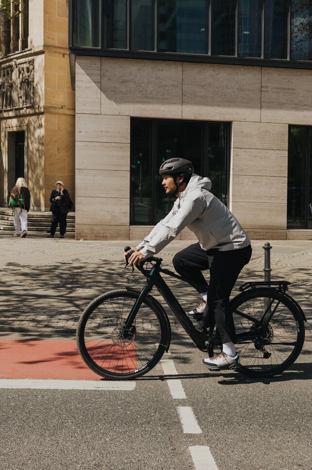 Ein junger Mann fährt in Freizeitkleidung und mit Fahrradhelm mit einem eBike auf einer Straße in der Stadt und überquert dabei einen roten Fahrradweg. Im Hintergrund sind moderne Gebäude und Passanten zu sehen.