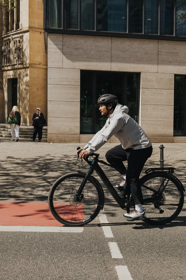 En ung man i vardagskläder och cykelhjälm cyklar elcykel på en stadsgata och korsar en röd cykelväg. I bakgrunden syns moderna byggnader och förbipasserande.