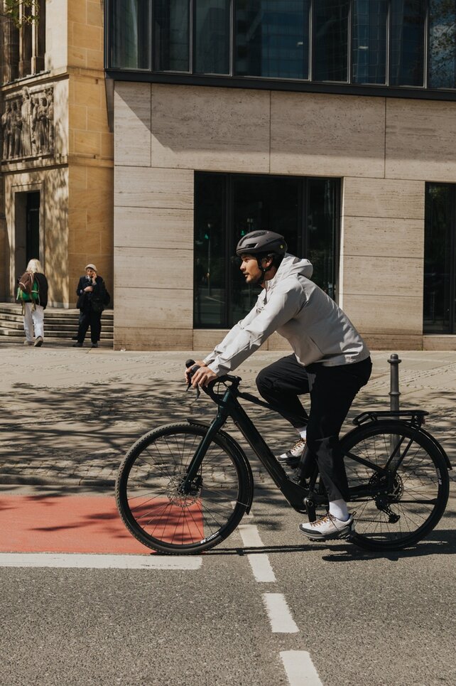 Ein junger Mann fährt in Freizeitkleidung und mit Fahrradhelm mit einem eBike auf einer Straße in der Stadt und überquert dabei einen roten Fahrradweg. Im Hintergrund sind moderne Gebäude und Passanten zu sehen.