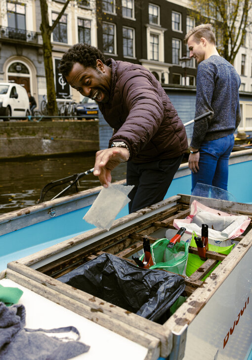 Een man op een kleine boot houdt een net vast en gooit met zijn andere hand plastic afval in een vuilnisbak op de boot. Hij kijkt tevreden.