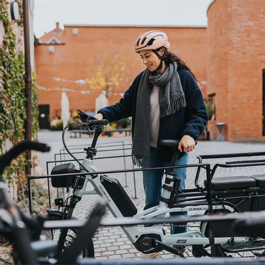 Woman with a pink helmet and gray scarf stands next to an eBike at the bike racks, smiling.
