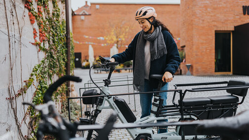 Frau mit rosanem Helm und grauem Schal steht neben eBike an Fahrradständern und lächelt.
