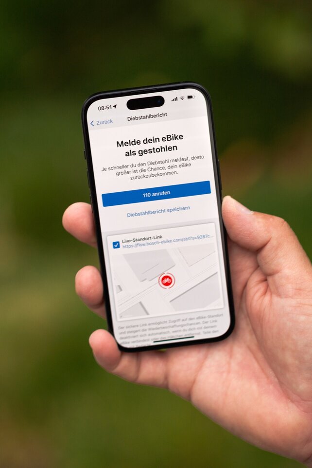 Eine Hand hält ein Smartphone, das den Bildschirm 'Melde dein eBike als gestohlen' der Bosch eBike Flow App anzeigt. Die App ermöglicht es, den Diebstahl zu melden, direkt die Polizei (110) anzurufen und einen Live-Standort-Link zur Nachverfolgung zu erstellen.