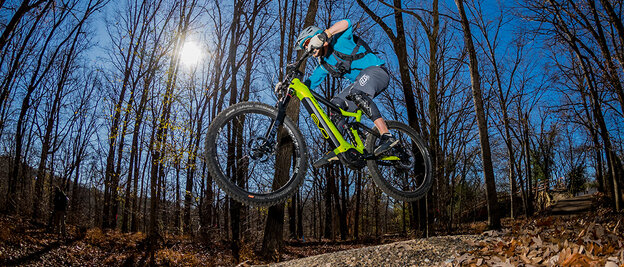 At dusk, an eMountain biker soars through the air along a trail in the forest. The moon shines through the bare trees, and leaves cover the ground.