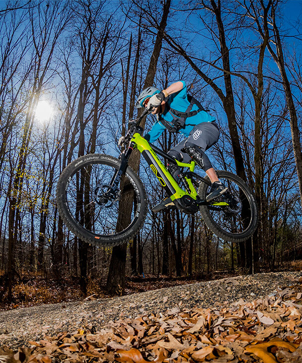 At dusk, an eMountain biker soars through the air along a trail in the forest. The moon shines through the bare trees, and leaves cover the ground.