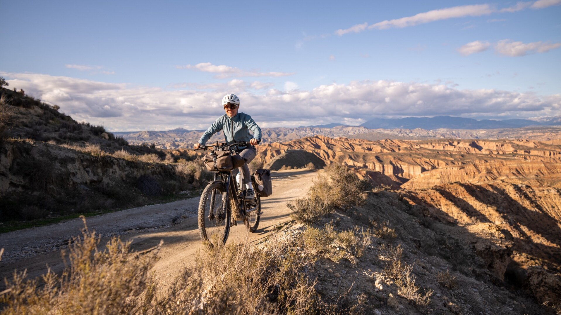 A person rides a loaded eBike along a dirt path on a mountain ridge, wearing a helmet and smiling at the camera.
