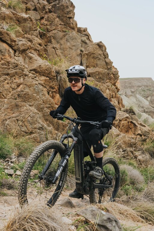 A mountain biker in full gear rides his eMTB on a technically demanding trail through a rocky, barren mountain landscape.