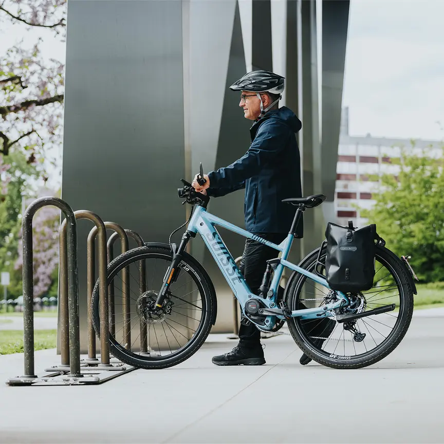 A person stands next to a blue e-bike, wearing a black helmet and a dark blue jacket. The bike is parked at a bicycle rack. In the background, you can see modern buildings and blooming trees.