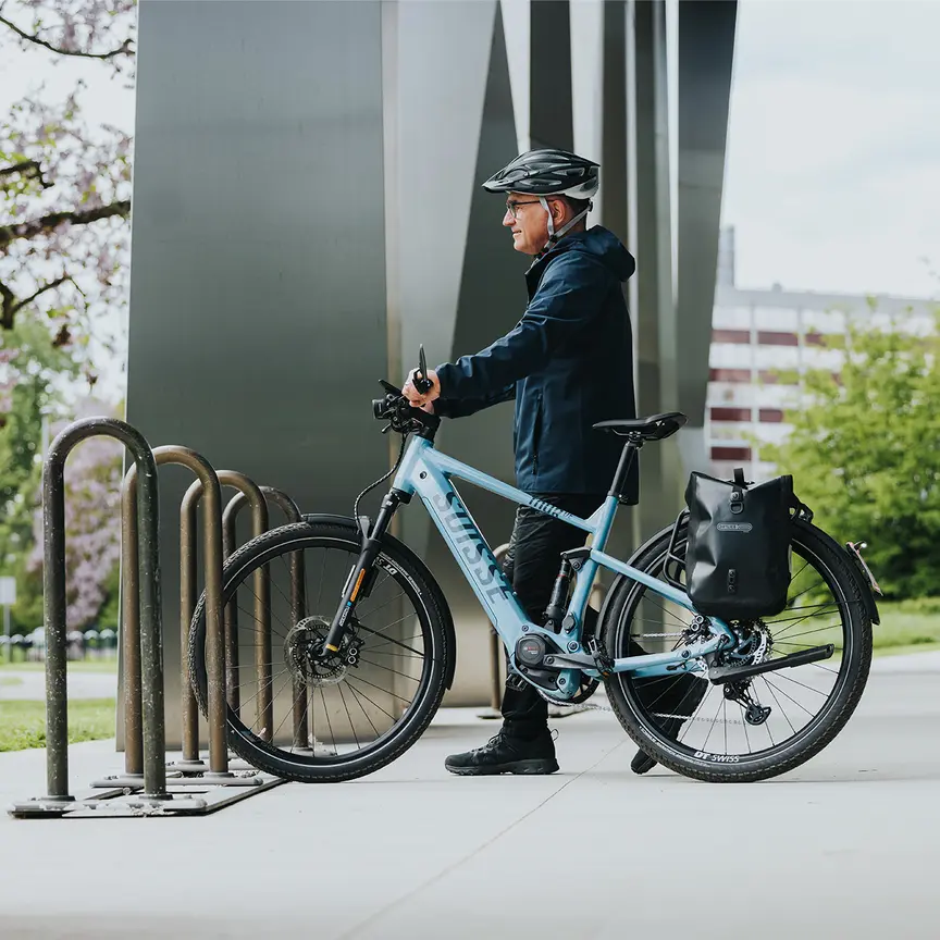 A person stands next to a blue e-bike, wearing a black helmet and a dark blue jacket. The bike is parked at a bicycle rack. In the background, you can see modern buildings and blooming trees.