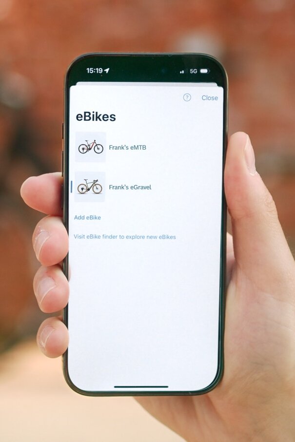Close-up of a smartphone showing the eBike Flow app's digital garage. The display screen lists the user's personal eBikes, including an eMTB and a Gravel eBike, with the option to add more eBikes.