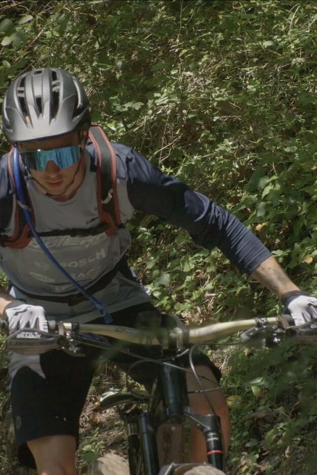 Vue de face d’un homme qui roule sur un VTT à assistance électrique sur un parcours très boisé. Il porte un casque, un sac à dos et des lunettes de soleil miroir.