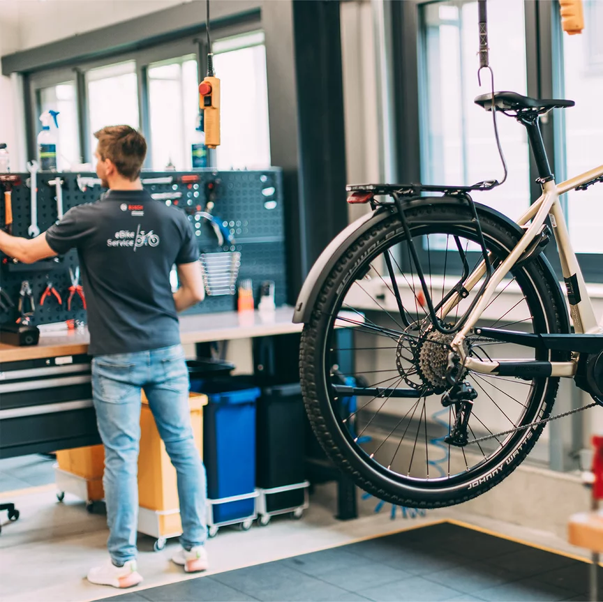Ein eBike hängt über dem Boden in einer Werkstatt. Im Hintergrund sieht man einen Bosch Mitarbeiter.