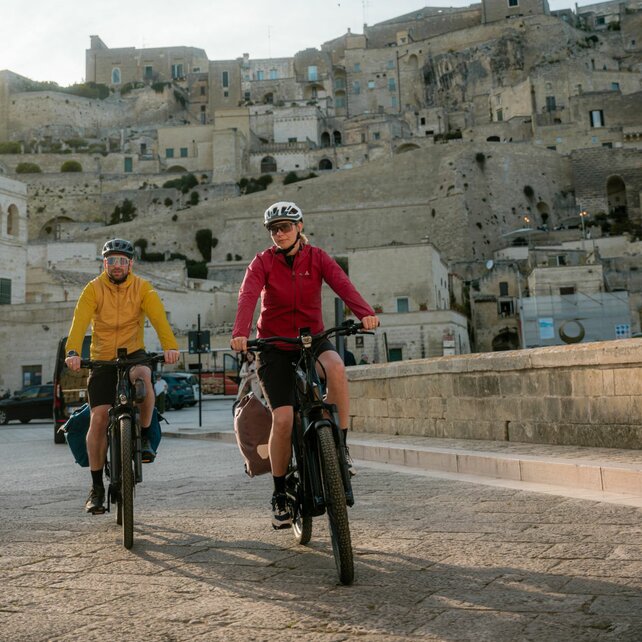 A woman and a man are riding their eBikes along a street with an Italian old town in the background.