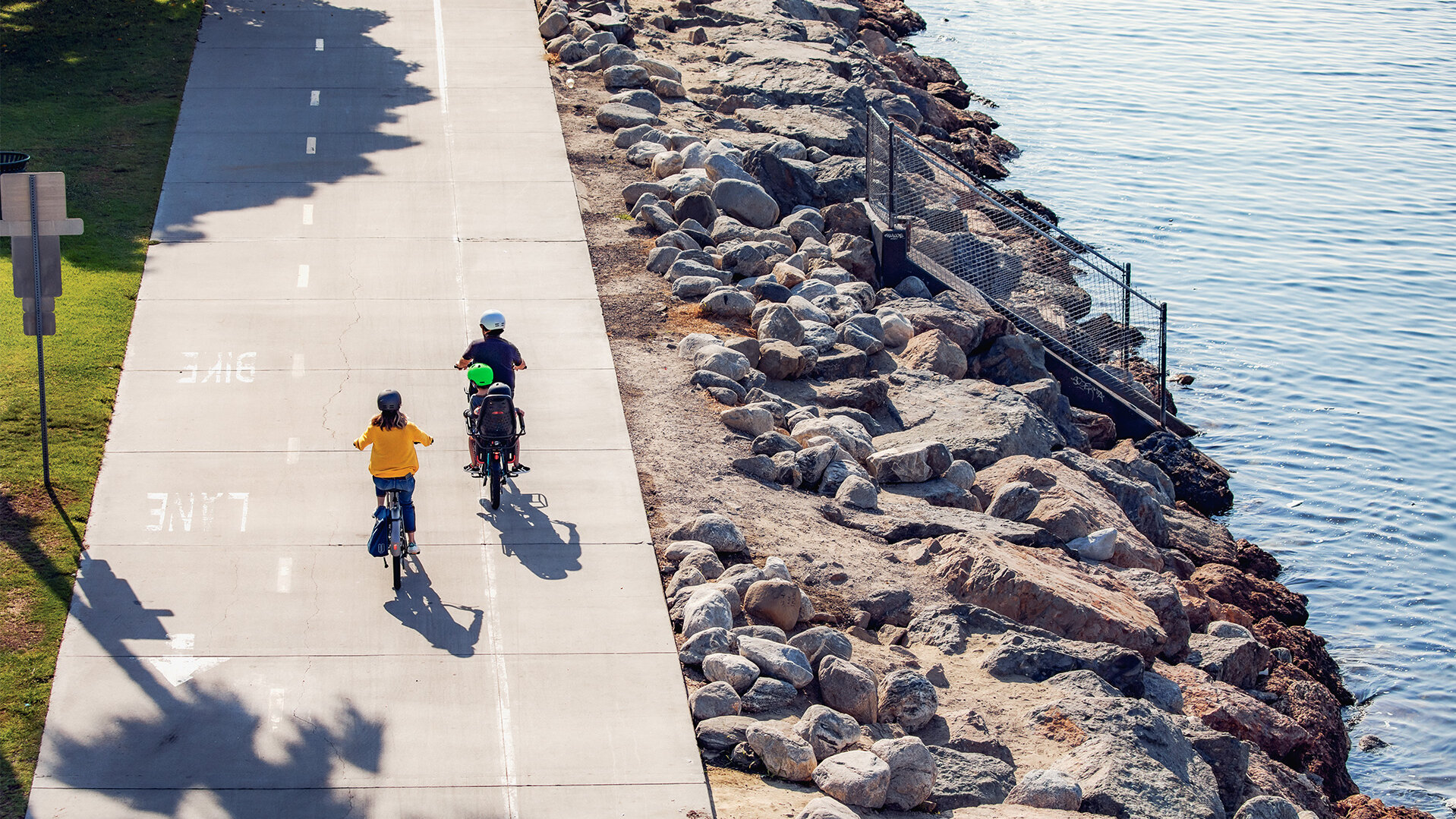 Two rider/eBike rider/eBiker riding on a bicycle road long a river. One of the rider/eBike rider/eBiker also has two children on the luggage rack.