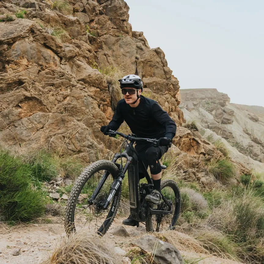 En eMountainbiker kører stejlt opad ad en smal sti gennem et barsk, klippefyldt landskab. I baggrunden strækker en bred, tør kløft sig ud under en lys himmel.